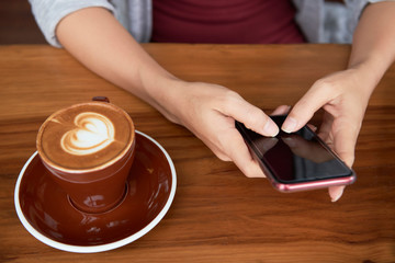 Fototapeta na wymiar Close-up of woman sitting at wooden table with cup of coffee and typing a message on her smartphone
