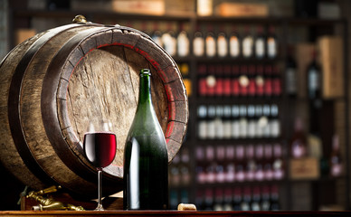 Wine bottle and glass of red wine on wooden cask. Wine shelves at the background.
