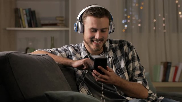 Happy man listening to music on smart phone sitting on a couch at home in the night