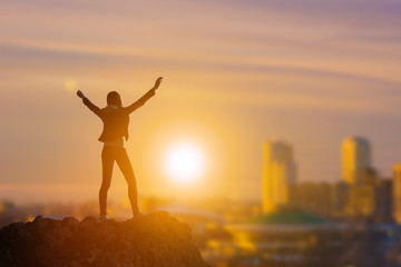 Fototapeta na wymiar Slender girl businessman on top of a mountain holding her hand up, against the background of the city in the rays of the sun. Business concept idea, happiness, success and achievement, leadership.
