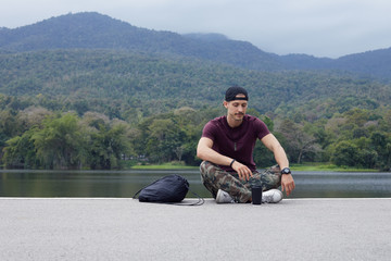 Attractive European hipster sitting on ground with lake view background, taking rest at city park...