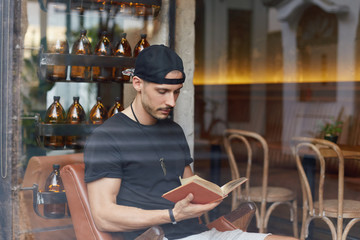 Handsome young man reading book in modern cafeteria with stylish interior design, looking concentrated, interested. Confident freelancer taking rest from gadgets with printed old literature in hands.