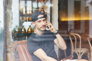 Portrait of attractive Caucasian man wearing sporty clothes, looking through window while listening to messages on his mobile phone, with thoughtful and calm face expression against decorated wall.