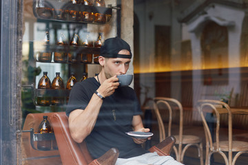 Portrait through window of young attractive bearded European man in casual clothes relaxing in stylish modern cafe indoors. Has concentrated and thoughtful look, making a sip of fresh brewed coffee.