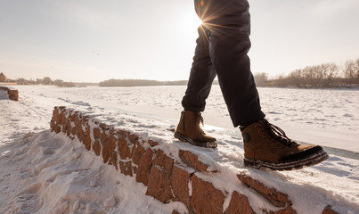 Wide angle view of man's legs walking along the fence on the shore of a frozen river covered with snow, scene lit by bright sun light reflected from snow.