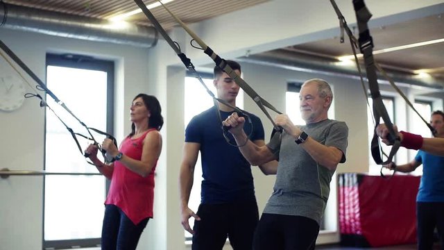 A group of cheerful seniors in gym with a young trainer doing exercise with TRX.