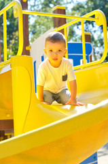 A cute two-year-old boy in a white t shirt sliding down on a yellow slide on a playground