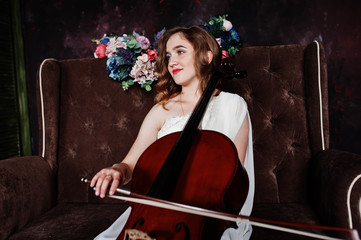 Pretty young gilrl musician in white dress with double bass sitting on brown vintage sofa.