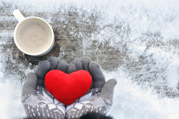 Valentine's day and Love frosty picture: a Cup of hot coffee on a wooden bench and hands in knitted gray gloves hold a plush red heart on a winter day. Valentine day