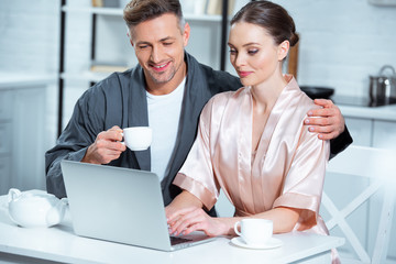 adult couple in robes sitting at table with tea cups and using laptop during breakfast in kitchen