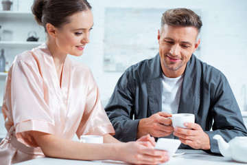 beautiful couple in robes using smartphone while having tea during breakfast in kitchen