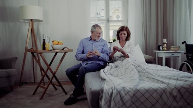 A senior couple sitting on bed at home, a man giving medication to ill wife.