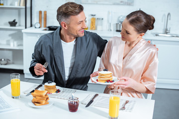 selective focus of adult couple in robes during breakfast with pancakes and orange juice in kitchen