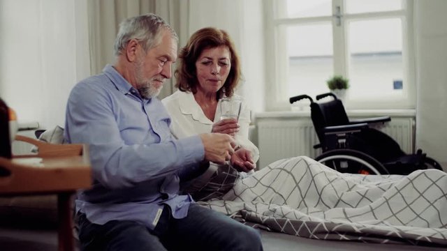 A senior couple sitting on bed at home, a man giving medication to ill wife.