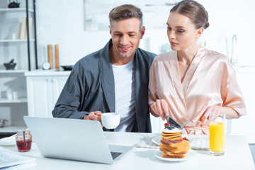 Fototapeta na wymiar adult couple in robes sitting at table with laptop while having breakfast in kitchen