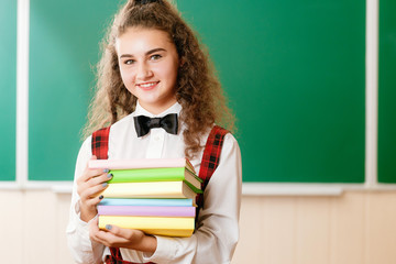 beautiful schoolgirl stands in the classroom and holds books against the background of the blackboard