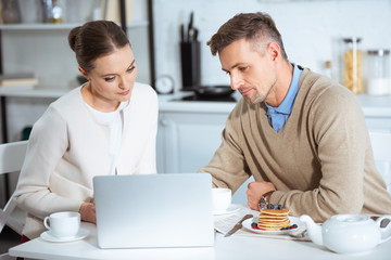 adult couple sitting at table and using laptop during breakfast in morning
