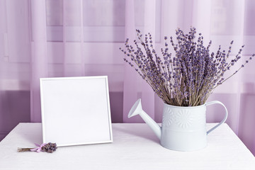Lavender flowers in watering can and photo frame (mock-up) on window with tulle cloth background, copy space.  Soft focus.