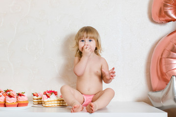 Little blonde baby girl two years old in pink pants sitting on the white table near her birthday cake and different pink sweets on the table