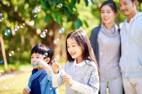 little asian boy and girl blowing bubbles outdoors parents watching from behind