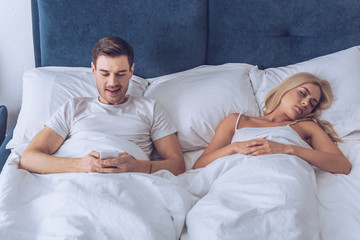 smiling young man lying in bed and using smartphone while young wife sleeping, secret concept