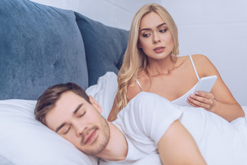 Obraz premium jealous woman holding smartphone and looking at husband sleeping in bed, mistrust concept