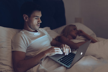 suspicious young man using laptop while girlfriend sleeping in bed at night