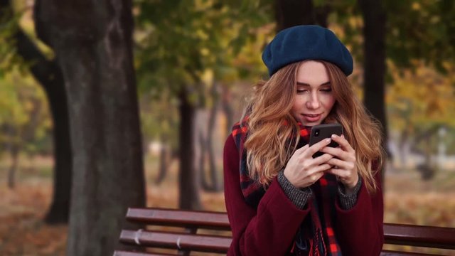 Smiling brunette woman in beret hat and coat writing message on smartphone while sitting on bench in park