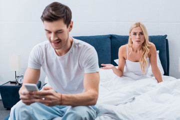 smiling young man in pajamas using smartphone and angry girlfriend looking at camera in bedroom, mistrust concept