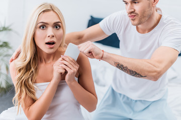 jealous man grabbing smartphone from scared girlfriend looking at camera in bedroom