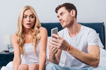 jealous man showing smartphone to confused girlfriend sitting on bed, distrust concept