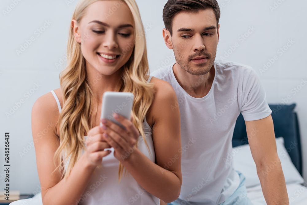 Wall mural jealous man looking at beautiful smiling wife using smartphone in bedroom, mistrust concept