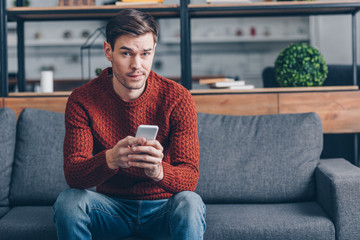 suspicious young man holding smartphone and looking at camera while sitting on couch at home - obrazy, fototapety, plakaty