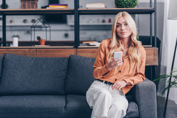 Obraz premium attractive blonde girl using smartphone and smiling at camera while sitting on couch at home