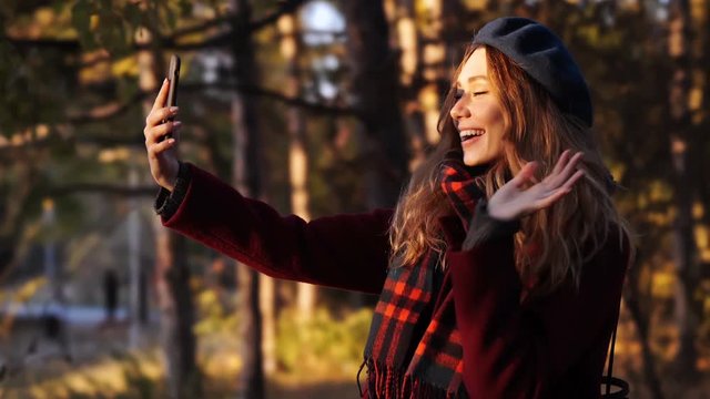 Happy brunette woman in beret hat and coat making selfie on smartphone while standing in park