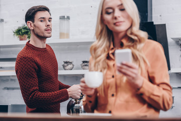 selective focus of jealous man looking at smiling girlfriend holding cup and using smartphone in kitchen
