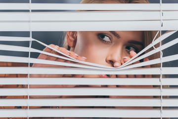young woman looking away and peeking through blinds, mistrust concept