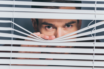 suspicious young man looking at camera through blinds, mistrust concept - obrazy, fototapety, plakaty