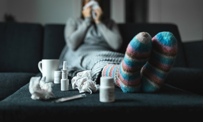 Woman sneezing and blowing nose with tissue and handkerchief. Sick and ill person with flu, cold medicine and woolen socks. Fever, virus or infection concept. Sitting on couch at home in winter. - obrazy, fototapety, plakaty