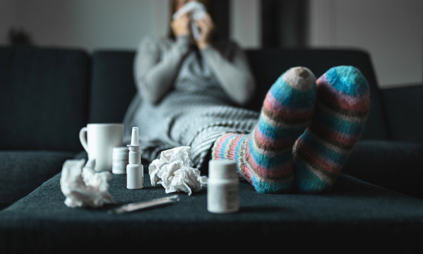 Woman sneezing and blowing nose with tissue and handkerchief. Sick and ill person with flu, cold medicine and woolen socks. Fever, virus or infection concept. Sitting on couch at home in winter.
