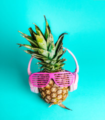 Fashionable trendy pineapple fruit with headphones and sun glasses listen to the music over bright pastel cyan background.