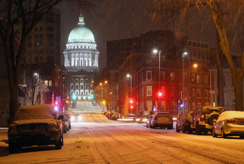 Snowy storm in a city background. Weather alert concept. Blizzard night downtown cityscape with glowing capitol building and baked cars along street. Midwest USA, Wisconsin, Madison.