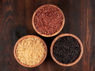 Uncooked black, red and integral rice in olive wood bowls, on a dark rustic wooden board, flat lay.