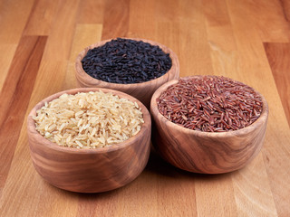 Uncooked black, red and integral rice in olive wood bowls, on a rustic wooden board.