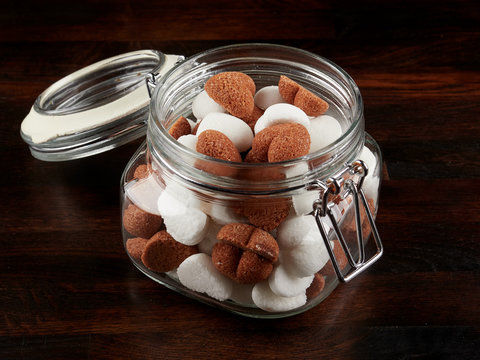 A jar filled sith white and brown heart shaped sugar cubes, on a dark wooden board