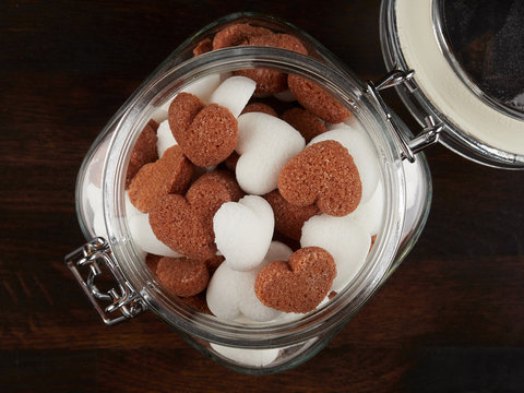 A jar filled sith white and brown heart shaped sugar cubes, on a dark wooden board, flat lay
