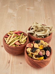Three olive wood bowls with uncooked, colorful orechiette, fusilli and tagliatelle pasta