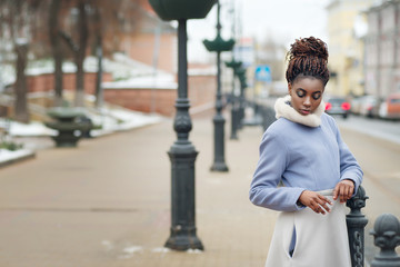 African girl in trendy coat and with makeup looks down on the city street on a winter day.
