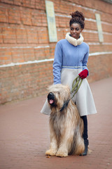 African girl in a stylish coat posing with a dog breed Briard on a city street on a winter day.