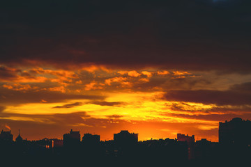 Cityscape with vivid fiery dawn. Amazing warm dramatic cloudy sky above dark silhouettes of city buildings. Orange sunlight. Atmospheric background of sunrise in overcast weather. Copy space.